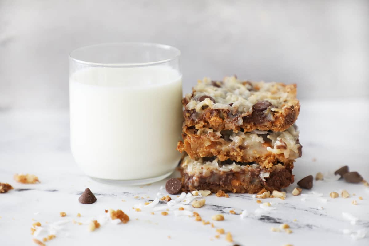 a glass of milk on a white marble counter next to a stack of magic cookie bars